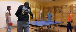 Stół do tenisa stołowego, wokół stołu stoją cztery osoby-trzech chłopców i jedna dziewczyna, w rękach trzymają paletki i grają mecz. Na ścianach widać drabinki gimnastyczne.