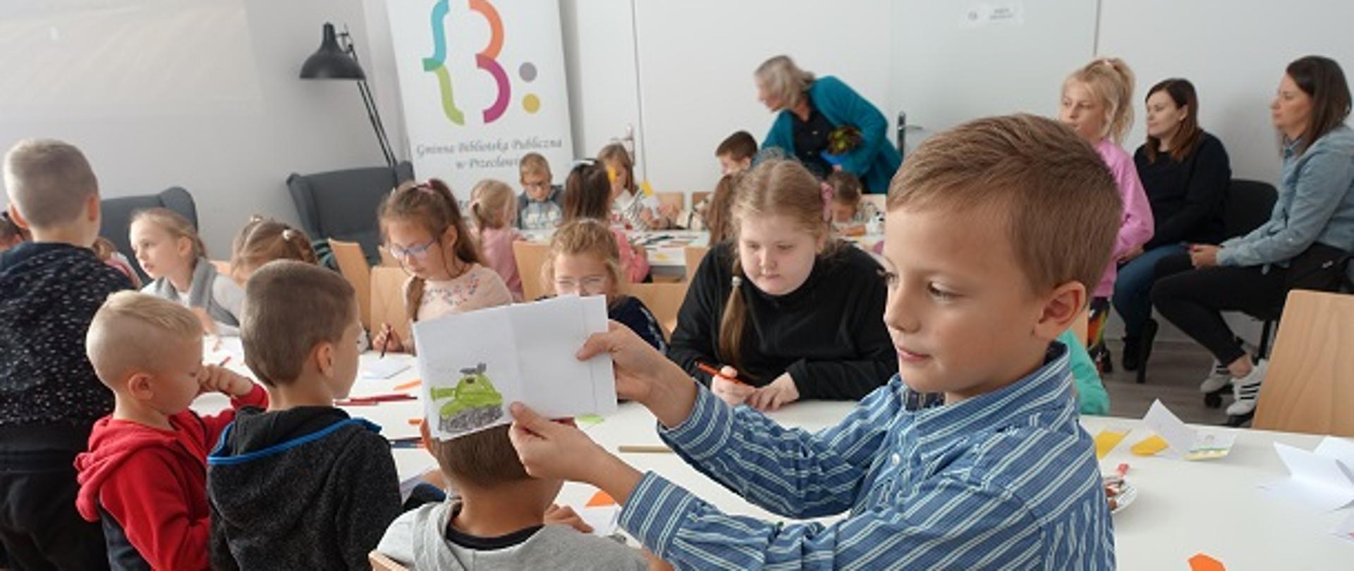Uczeń prezentuje wykonaną przez siebie książeczkę. W tle przy stolikach pracują uczniowie.