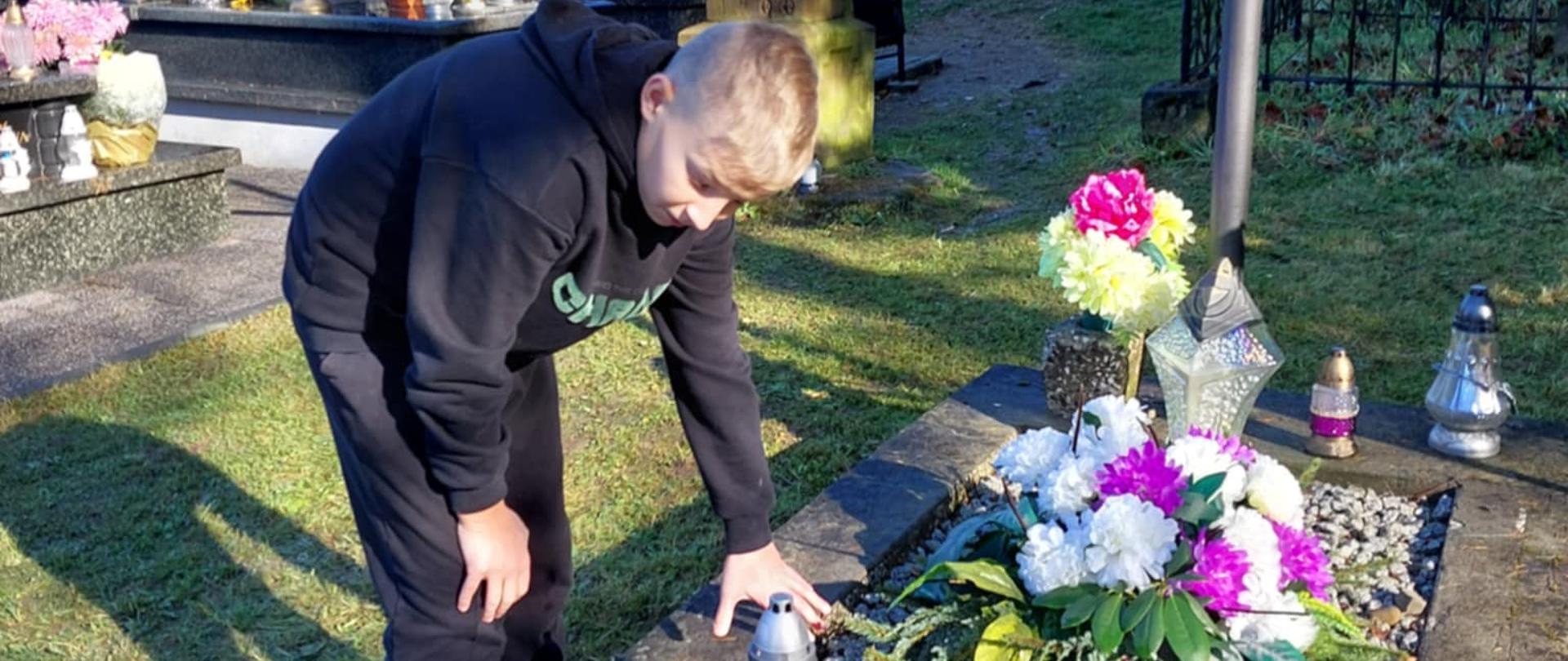Uczeń pochyla się nad grobem, żeby zapalić znicz, w tle widoczne inne groby