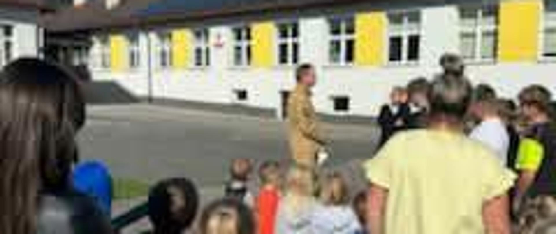 Na zdjęciu widać grupę uczniów zgromadzonych przed budynkiem szkoły. Dzieci stoją w niewielkich grupach i skupiają uwagę na osobach prowadzących spotkanie. W tle znajduje się szkolny budynek z charakterystycznymi żółtymi okiennicami oraz panelami fotowoltaicznymi na dachu. Całość odbywa się na szkolnym placu, w słoneczny dzień.