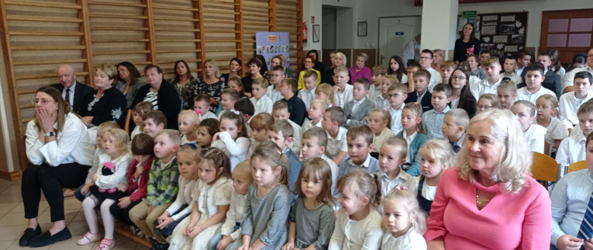 Uczniowie i nauczyciele siedzą na korytarzu szkolnym.