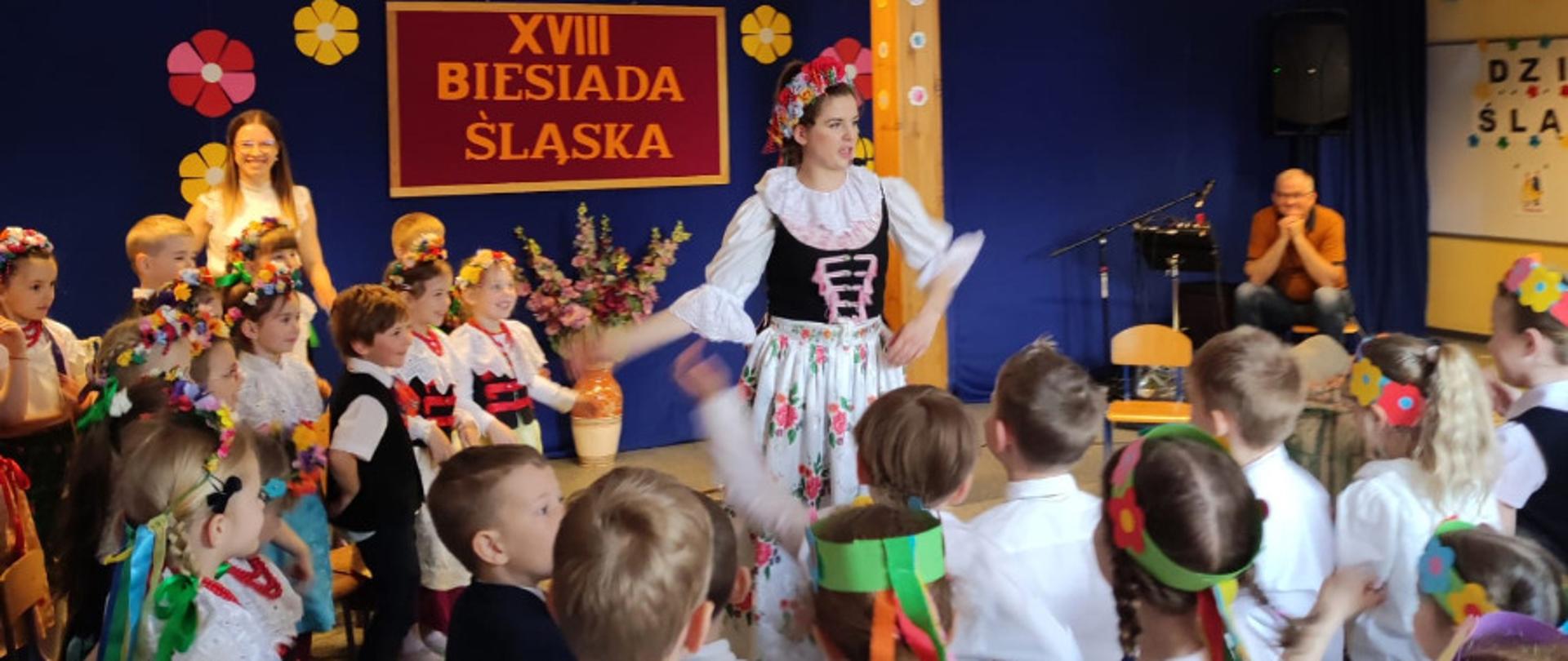 Kobieta w stroju śląskim wśród grupy przedszkolaków w tle napis XVIII biesiada śląska
