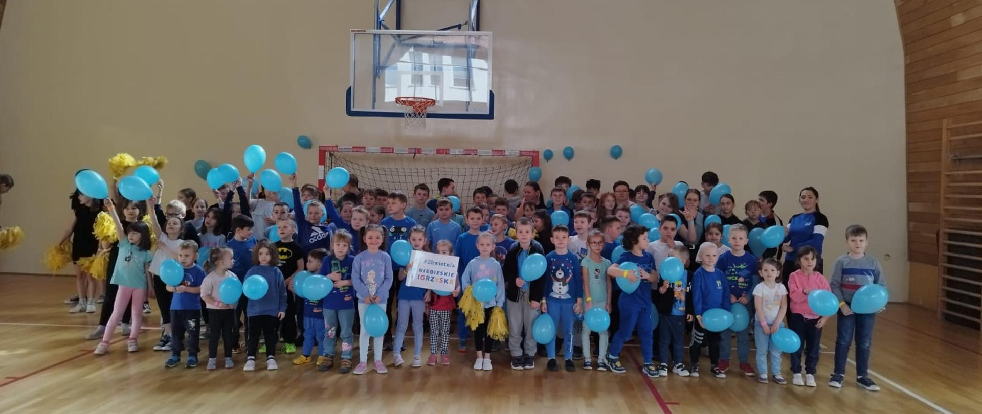 Wszyscy uczniowie szkoły ubrani na niebiesko, trzymając niebieskie balony stoją na sali gimnastycznej solidaryzując się tym gestem z osobami będącymi w spektrum autyzmu