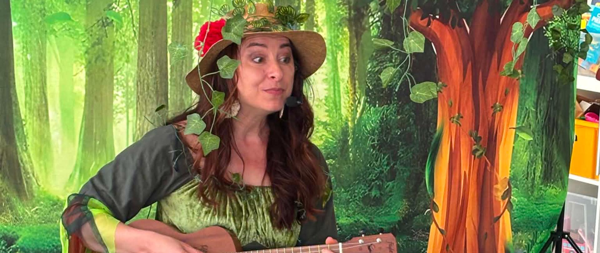Kobieta siedząca na krześle, w dłoniach trzyma ukulele. Jest ubrana w długą suknię khaki, a na głowie ma kapelusz. Za nią widać tło fotograficzne przedstawiające drzewa i zielone tło. Przed kobietą na ziemi leży walizka, obok stoi stojak na nuty.