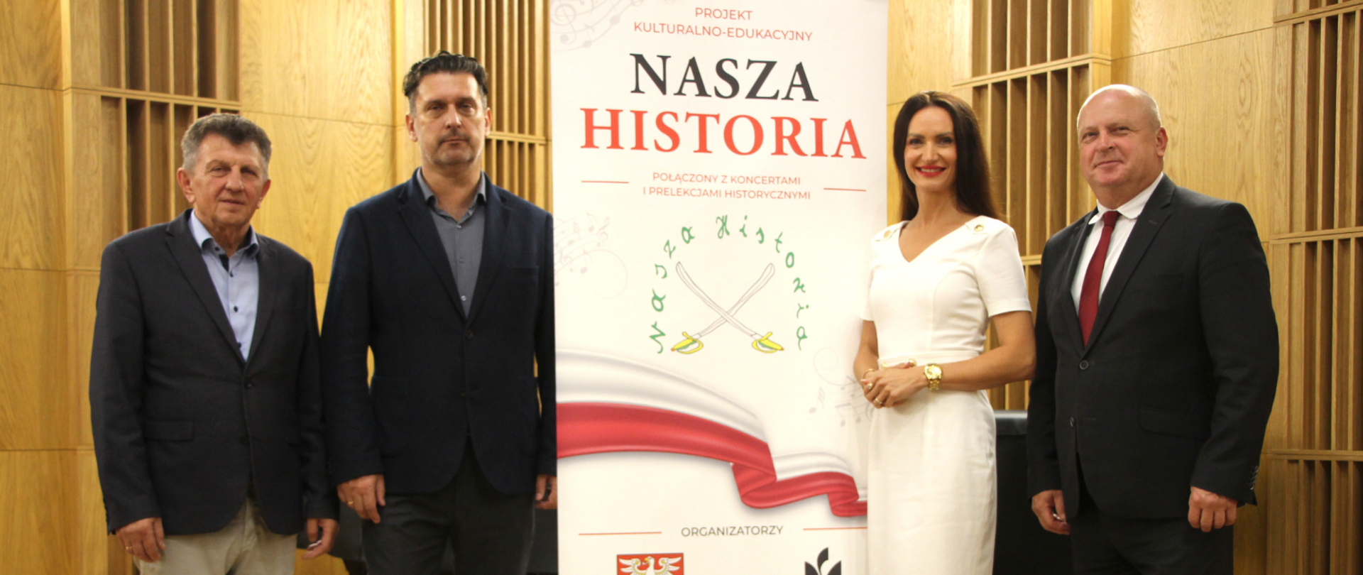 Przedstawia czworo organizatorów pozujących przy banerze projektu kulturalno-edukacyjnego „Nasza Historia”. W centrum uwagi znajduje się kobieta w eleganckiej białej sukience w otoczeniu trzech mężczyzn w garniturach.