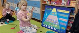 Dzieci z wykorzystaniem planszy edukacyjnej prezentują prowadzącej zajęcia jej poszczególne elementy
