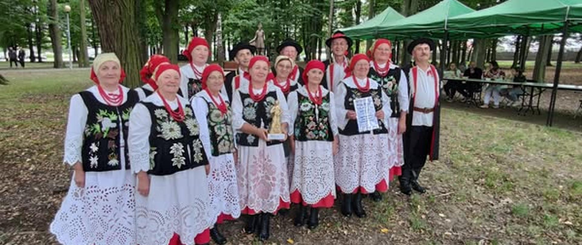 Kobiety w czarnych gorsetach i biało czerwonych spódnicach, w chustkach na głowie, mężczyźni w czarnych kapeluszach, białych koszulach i czarnych spodniach i płaszczach pozują do zdjęcia na tle zielonych drzew i namiotów 