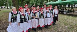 Kobiety w czarnych gorsetach i biało czerwonych spódnicach, w chustkach na głowie, mężczyźni w czarnych kapeluszach, białych koszulach i czarnych spodniach i płaszczach pozują do zdjęcia na tle zielonych drzew i namiotów