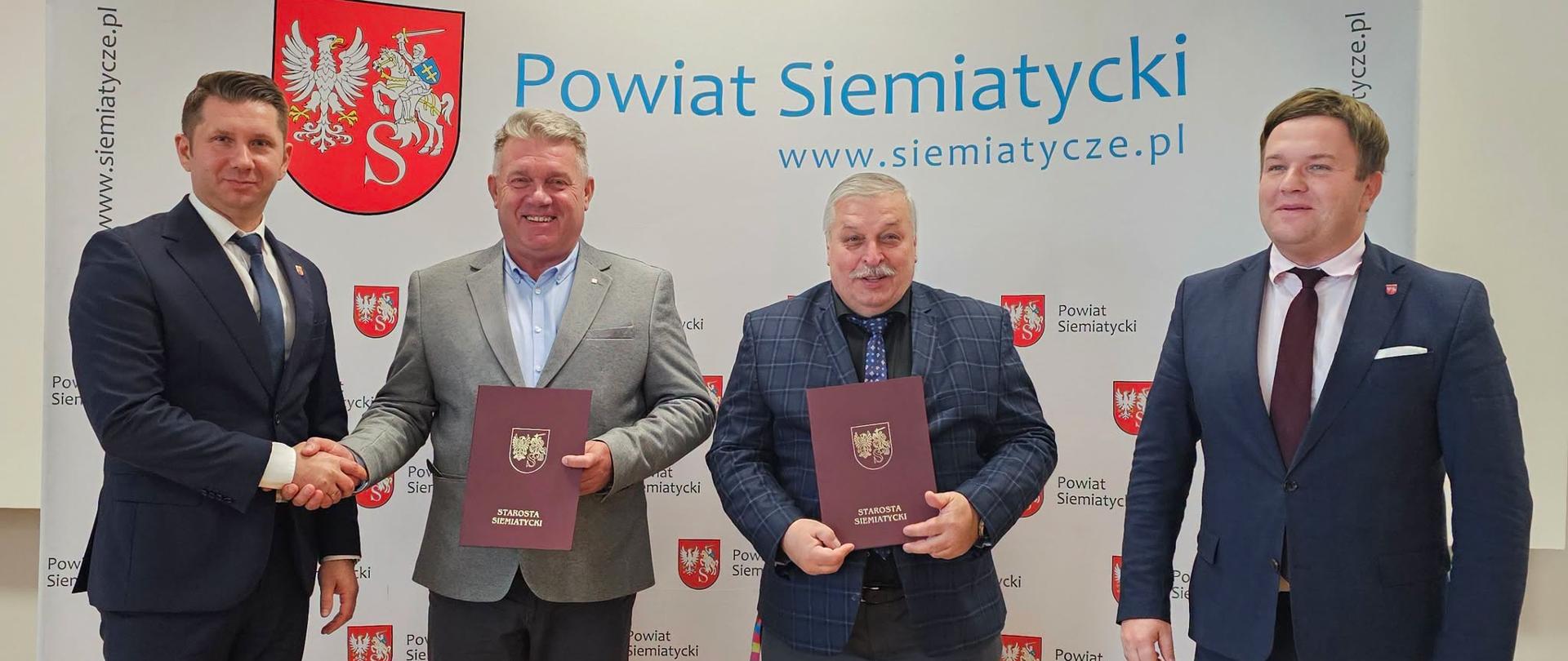 Czterech mężczyzn stoi przed transparentem z napisem „Powiat Siemiatycki”. Trzymają w rękach teczki i wygląda na to, że uczestniczą w uroczystym podpisaniu umowy lub ceremonii podpisania dokumentu.