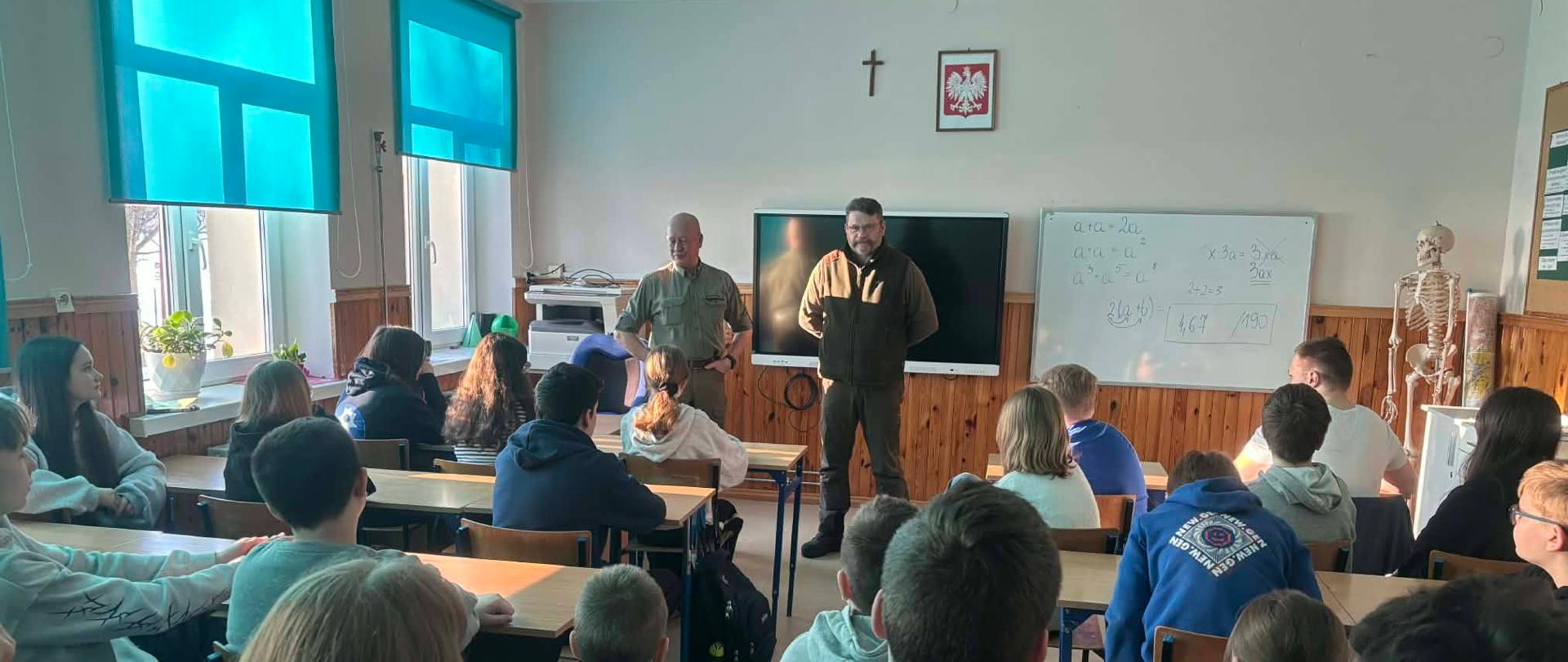 Uczniowie siedzą przy stolikach w sali lekcyjnej . Przed nimi stoi dwóch Panów w służbowych mundurach - przedstawicieli Nadleśnictwa w Augustowie.