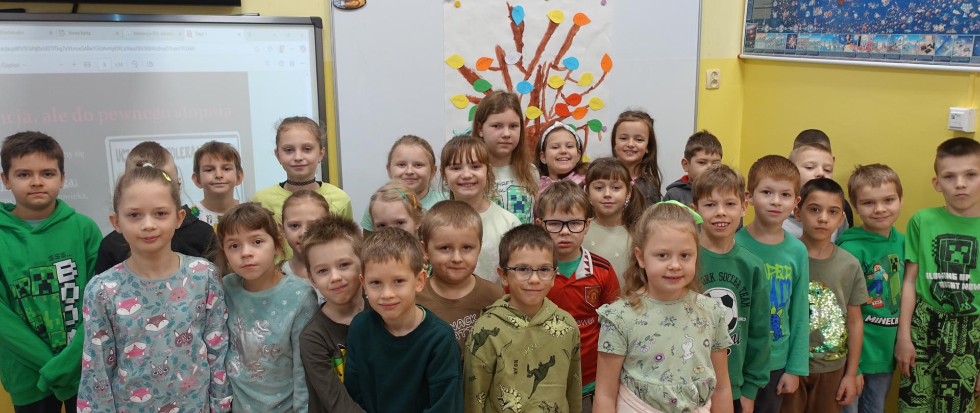 Uczniowie klas I-III stoja w sali lekcyjnej na tle szablonu drzewa
