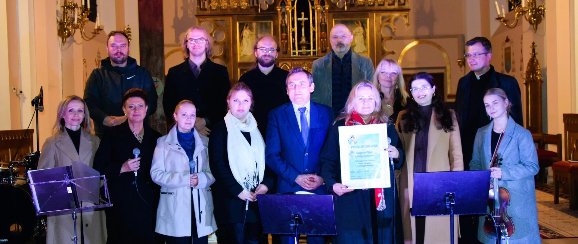 Grupa osób stoi wewnątrz kościoła przed ołtarzem, na tle dużego krzyża i złotych dekoracji. Niektórzy trzymają mikrofony, a jedna osoba prezentuje duży dyplom lub podziękowanie. Wokół widać instrumenty muzyczne i pulpity nutowe, co sugeruje, że odbywa się uroczyste wydarzenie lub koncert.
