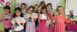 Grupa dziewczynek pozuje do zdjęcia z okazji święta Dnia Kobiet w przedszkolu. Dziewczynki trzymają w rękach upominki oraz dyplomy.