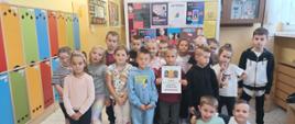 Dzieci z oddziału przedszkolnego ułożone w dwa rzędy stoją przez gazetką informacyjną dotyczącą zapobiegania przemocy, chłopiec na środku trzyma w ręku plakat z napisem "Tydzień o Przeciwdziałaniu Przemocy Rówieśniczej, dwóch chłopców siedzi na podłodze