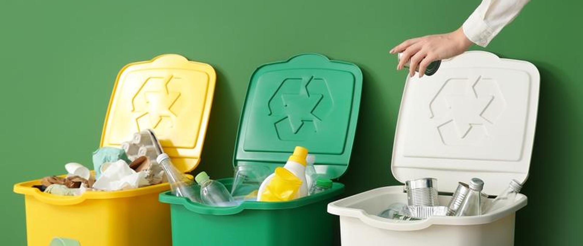 Zdjęcie przedstawia trzy pojemniki na odpady w kolorze żółtym, zielonym i białym, oraz dłoń wrzucającą odpady do pojemnika. 