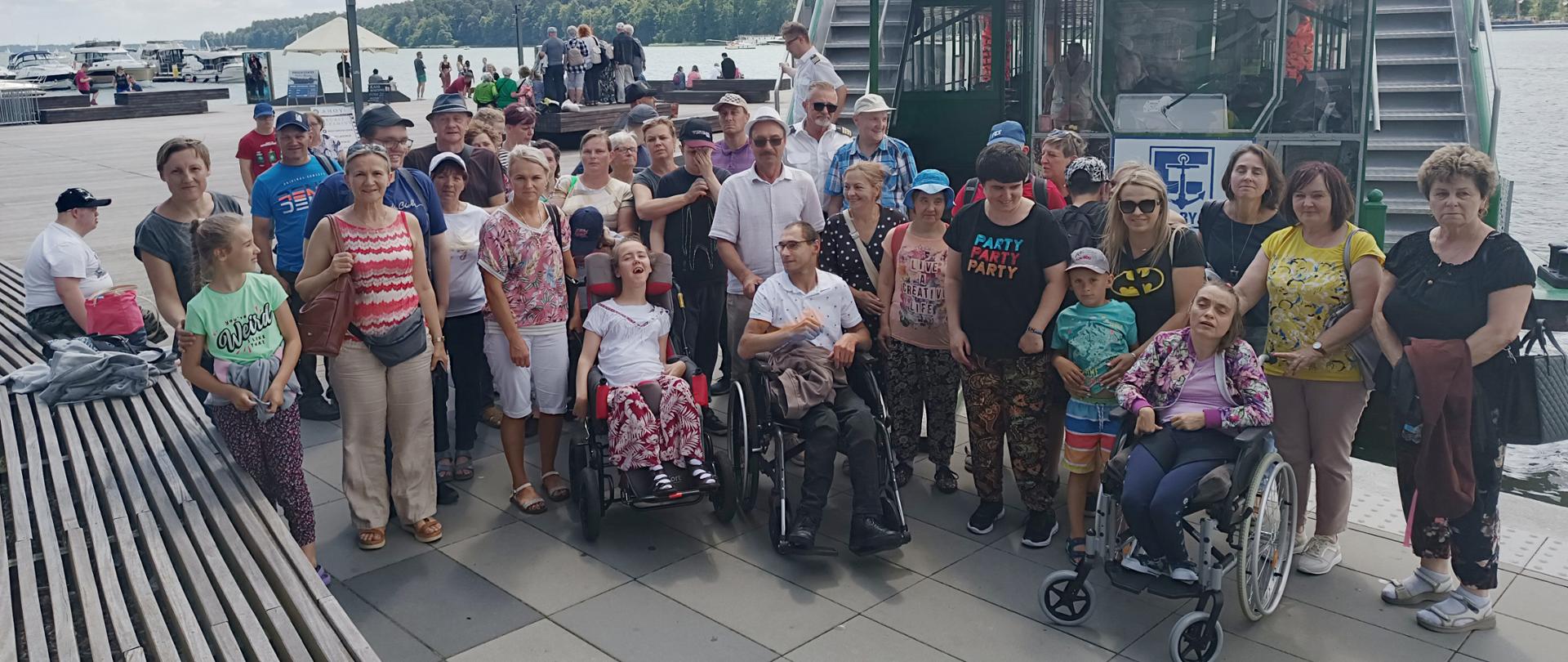 Grupa grupa osób dorosłych i dzieci z niepełnosprawnościami wraz z opiekunami na tle statku pasażerskiego i jeziora.