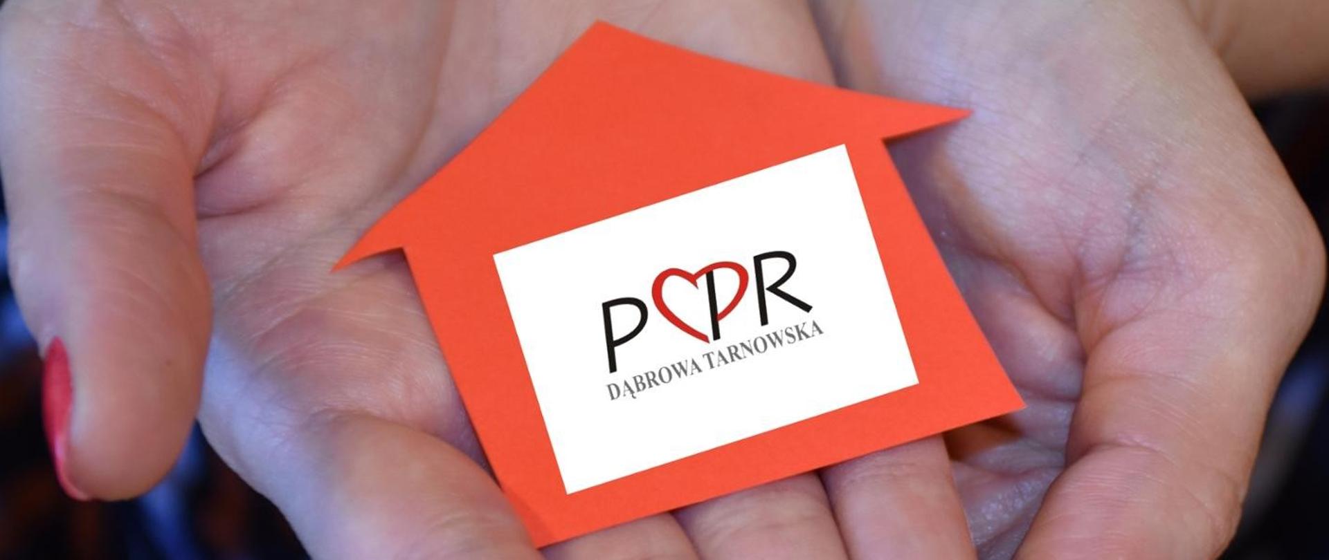 Dłonie kobiety trzymają wycięty z papieru pomarańczowy domek z białą naklejką i logo PCPR Dąbrowa Tarnowska