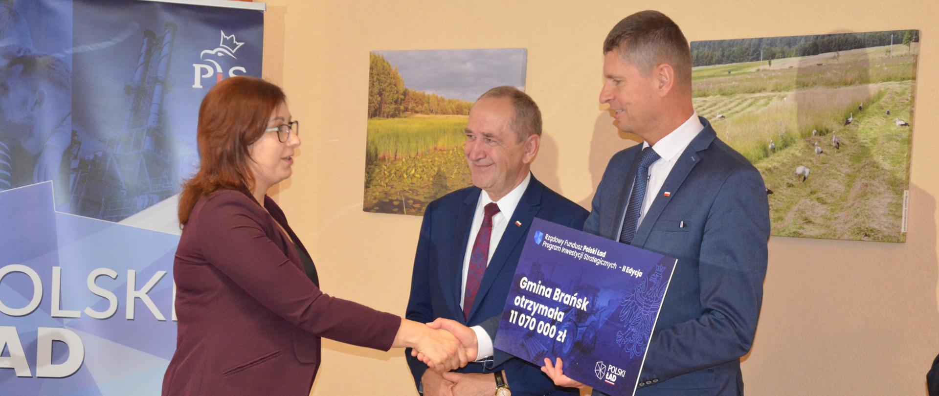 Kobieta w ciemnym ubraniu odbiera czak z rąk mężczyzny w garniturze.