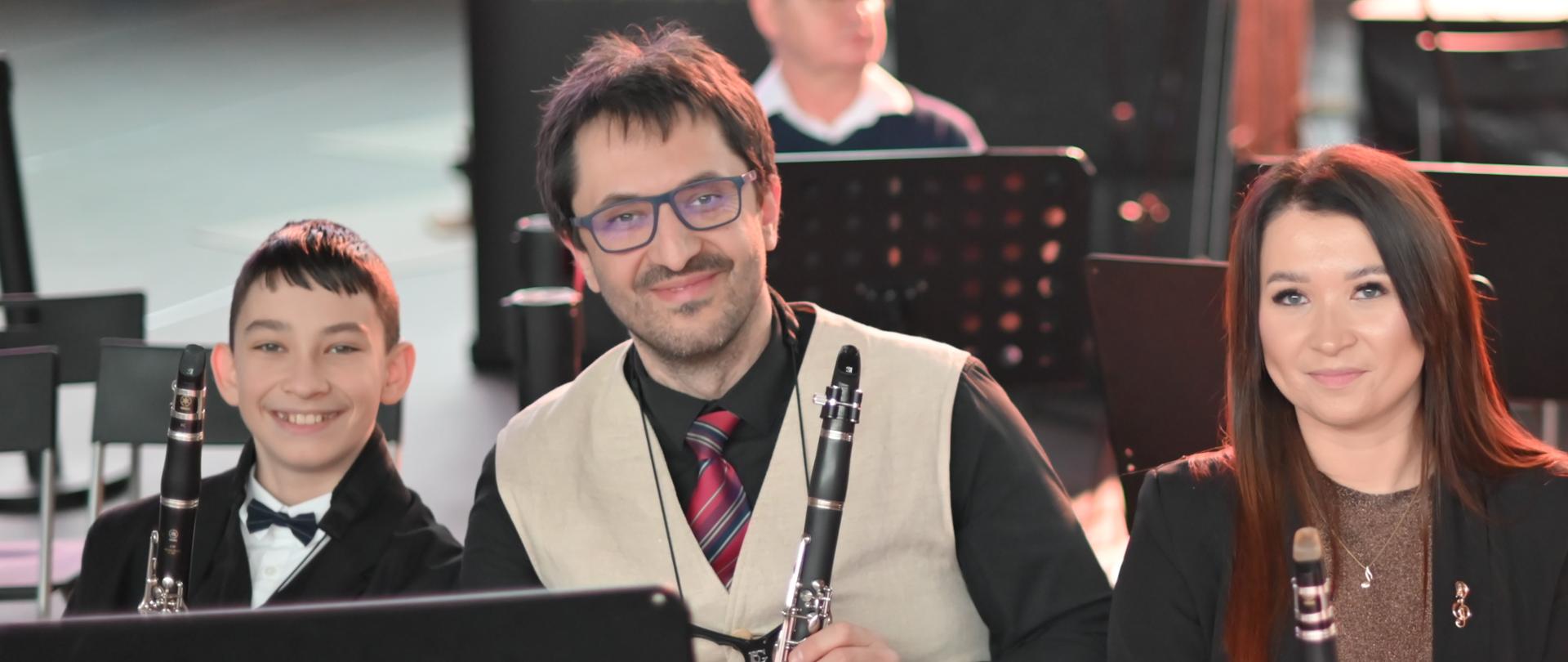 Troje muzyków orkiestry z klarnetami siedzi na estradzie i uśmiecha się do obiektywu.