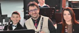 Troje muzyków orkiestry z klarnetami siedzi na estradzie i uśmiecha się do obiektywu.