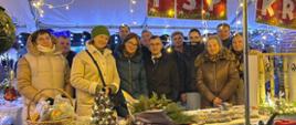 Grupa jedenastu osób uśmiechających się do kamery, stojących za stołem z bożonarodzeniowymi produktami pod białym namiotem. Wszyscy są ubrani w zimowe kurtki i płaszcze, a jedna z kobiet ma na głowie zieloną czapkę. Na stole, przykrytym częściowo białym, a częściowo bordowym obrusem, wyeksponowano wiklinowy kosz, małą choinkę z szyszek oraz stroiki z igliwia. Nad grupą wiszą sznury ciepłych, żółtych lampek oraz czerwone tabliczki z białymi literami. W tle widoczne są inne oświetlone stoiska, co tworzy nastrojową atmosferę wieczornego kiermaszu.