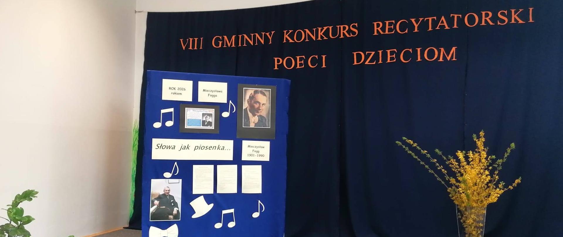 Na ciemej kurtynie wisi pomarańczowy napis VIII Konkurs Recytatorski Poeci Dzieciom. Z lewej strony stoi gazetka poświęcona Mieczysławowi Fogowi. Z prawej strony stoi wielki wazon z gałązkami złotego deszczyku.