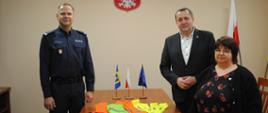 Wójt Gminy Raba Wyżna - Tomasz Rajca, Kierownik Gminnego Ośrodka Pomocy Społecznej- Katarzyna Kania oraz Komendant Powiatowej Policji w Nowym Targu- insp. Grzegorz Wyskiel w momencie przekazania gadżetów odblaskowych. Na stole leżą odblaski, w tle Godło Polski i flaga Polski.