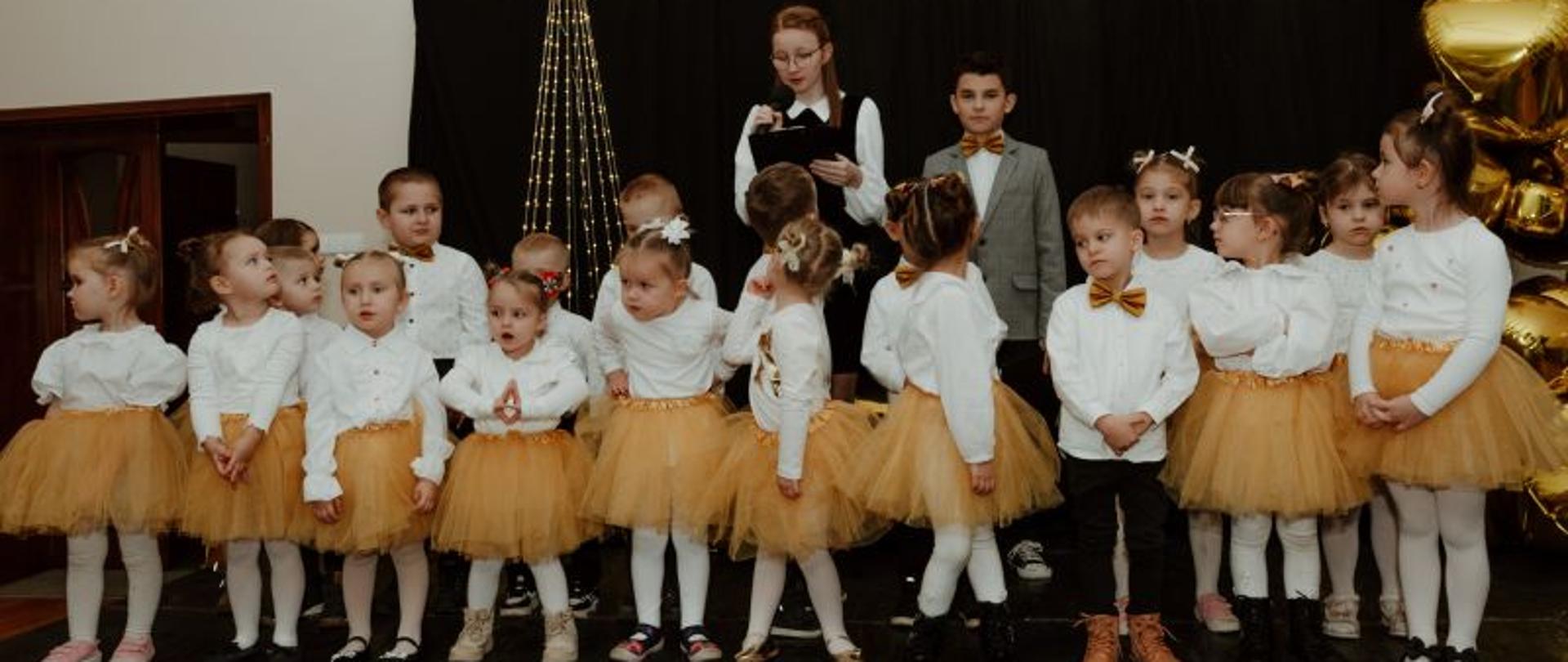Na scenie stoją dzieci z grupy Tygryski, za nimi uczniowie prowadzący koncert. 