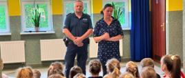 Policjant i pani pedagog rozpoczynają pogadankę na temat bezpiecznych wakacji, przed nimi na ławeczkach siedzą uczniowie