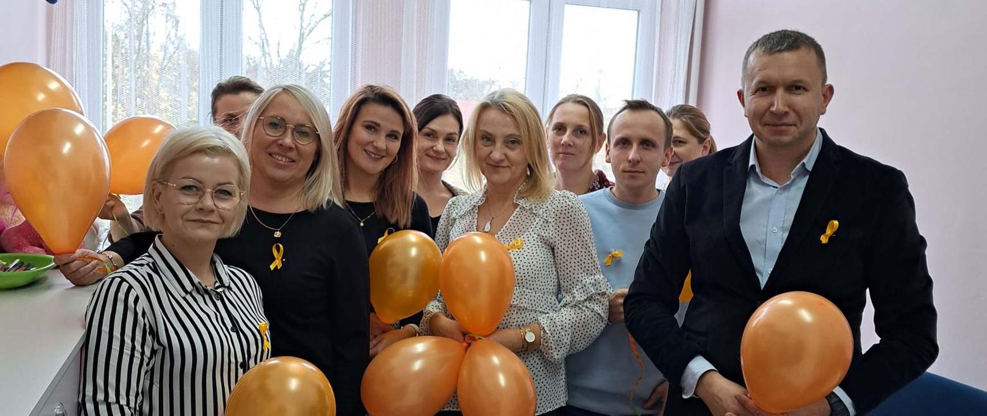 Zdjęcie przedstawia pracowników Powiatowego Centrum Pomocy Rodzinie w Przasnyszu promujących kampanię 19 dni przeciwko przemocy i krzywdzeniu dzieci i młodzieży - przypinając do ubrań pomarańczową wstążkę, udzielając informacji o akcji oraz rozdając pomarańczowe balony.