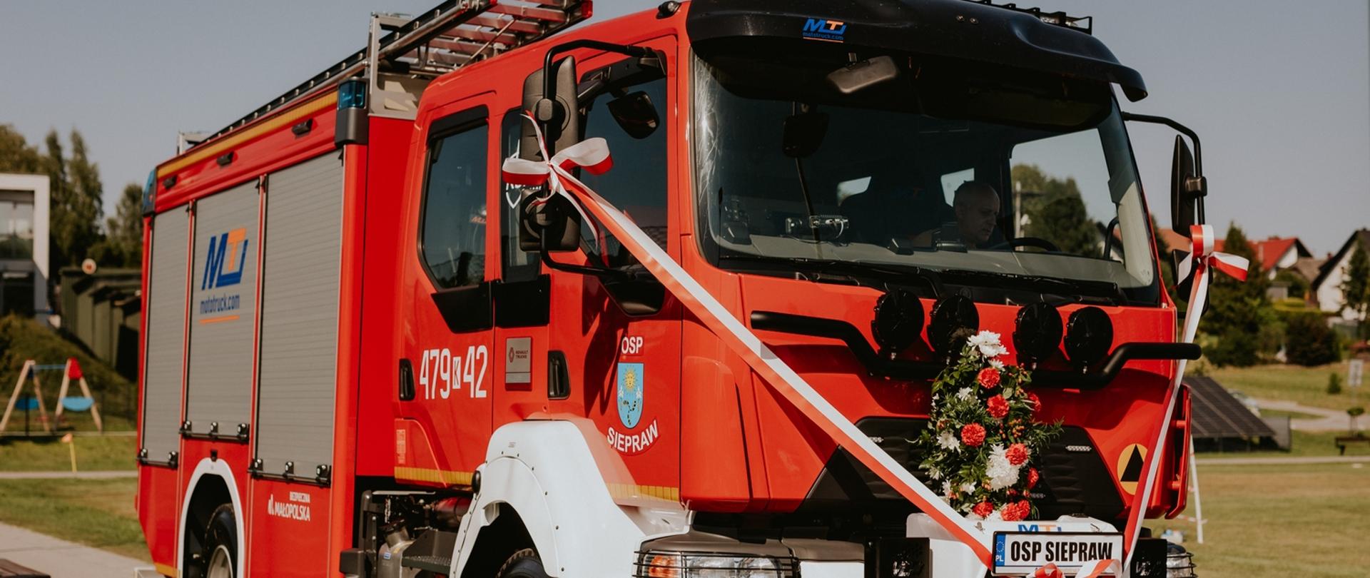 Czerwony wóz strażacki OSP Siepraw ozdobiony biało-czerwoną wstęgą i kwiatami stoi na trawie