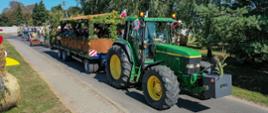 Korowód dożynkowy, z przodu traktor ciągnący zadaszoną przyczepę udekorowaną zbożem oraz kwiatami, w przyczepie na ławkach siedzą zaroszeni goście, za nimi jadą kolejne traktory ze sprzętem rolniczym