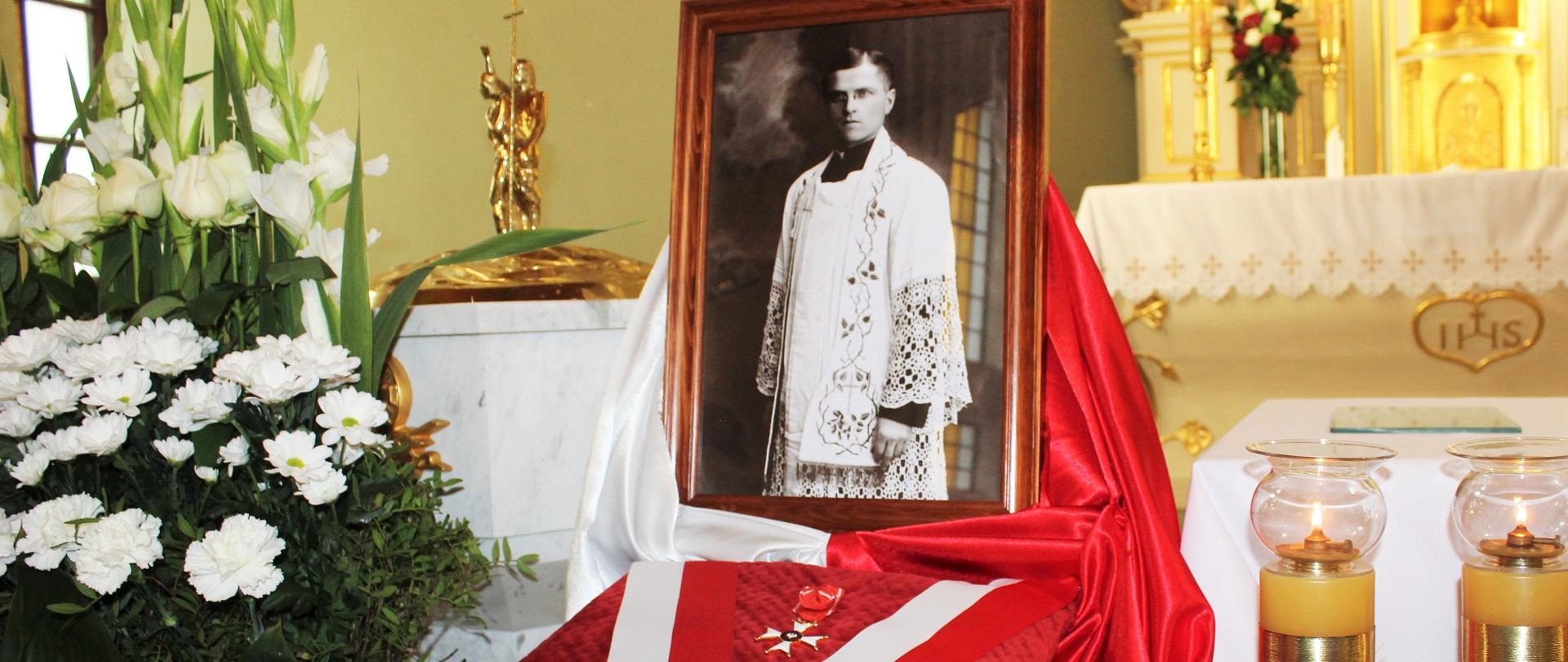 Na fotografii widoczne biało - czerwone kwiaty oraz czerwoną poduszkę przepasaną biało - czerwoną szarfą na której leży Krzyż Oficerski Orderu Odrodzenia Polski. Nad Poduszą ustawiony portret pośmiertnie odznaczonego ks. A. Łapaya. W drugim planie w tle ołtarz kościoła św. Wojciecha w Skibniewie.