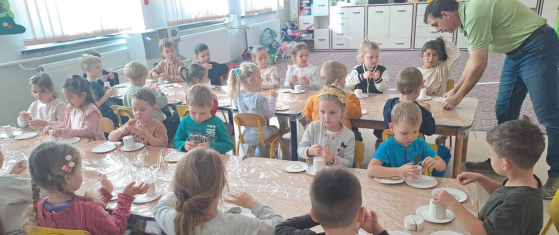 W sali przedszkolnej, w dwóch rzędach ustawione stoliki, przy których siedzą dzieci i wykonują prace manualne z wosku pszczelego. Przy nich stoi pan pszczelarz i coś objaśnia.