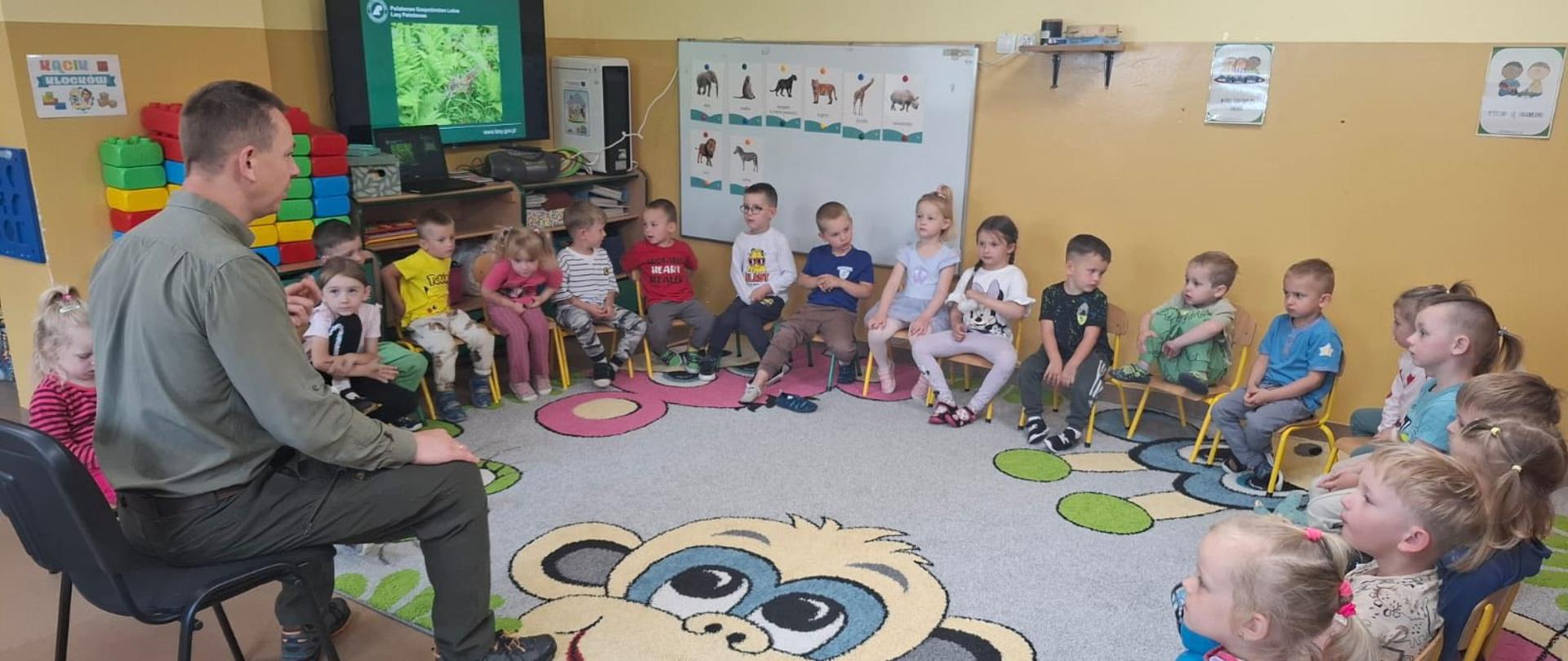 Dzieci przedszkolne siedzą na krzesełkach w kole, przed nimi pan leśnik wygłasza pogadankę, na ekranie wyświetla prezentację na temat lasu
