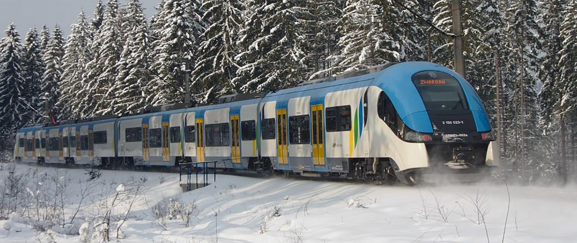 Biało-niebieski pociąg jadący po zaśnieżonych torach w tle pokryte śniegiem iglaste drzewa