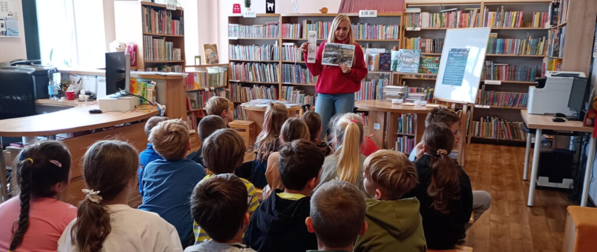 Zdjęcie przedstawia grupę dzieci siedzących na ławkach w bibliotece. Pani prowadząca warsztaty pokazuje zdjęcie gdzie zimują sarny. 