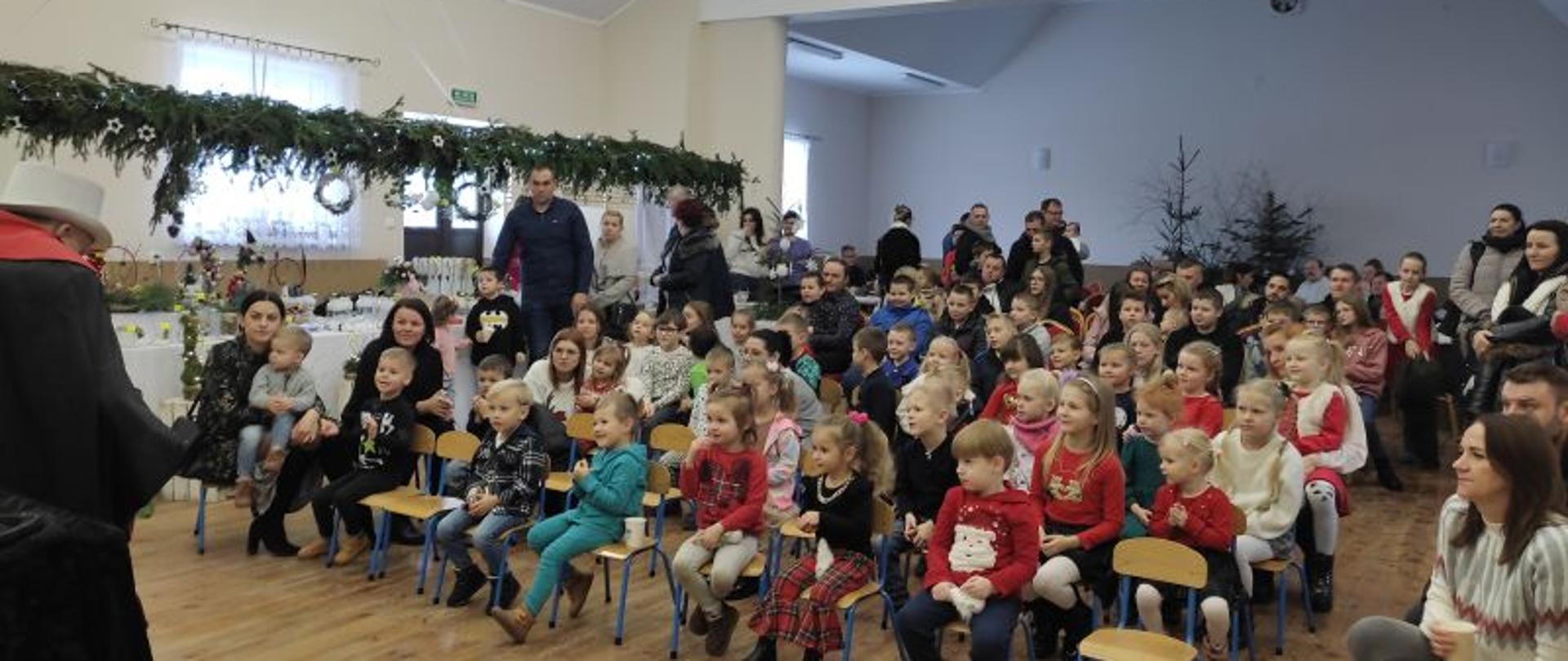 Duża grupa dzieci oraz dorośli siedzą na krzesłach, niektórzy stoją. Wszyscy przyglądają się magikowi w białym kapeluszu. 