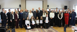 W centrum auli Centrum Edukacji Nauczycieli w Łomży stoi kilkudziesięciu laureatów, finalistów oraz gości, w tym Starosta Zambrowski Edyta Marchelska-Groszfeld. Osoby w pierwszym rzędzie prezentują otrzymane dyplomy i podziękowania. W tle widoczne jest godło Polski.