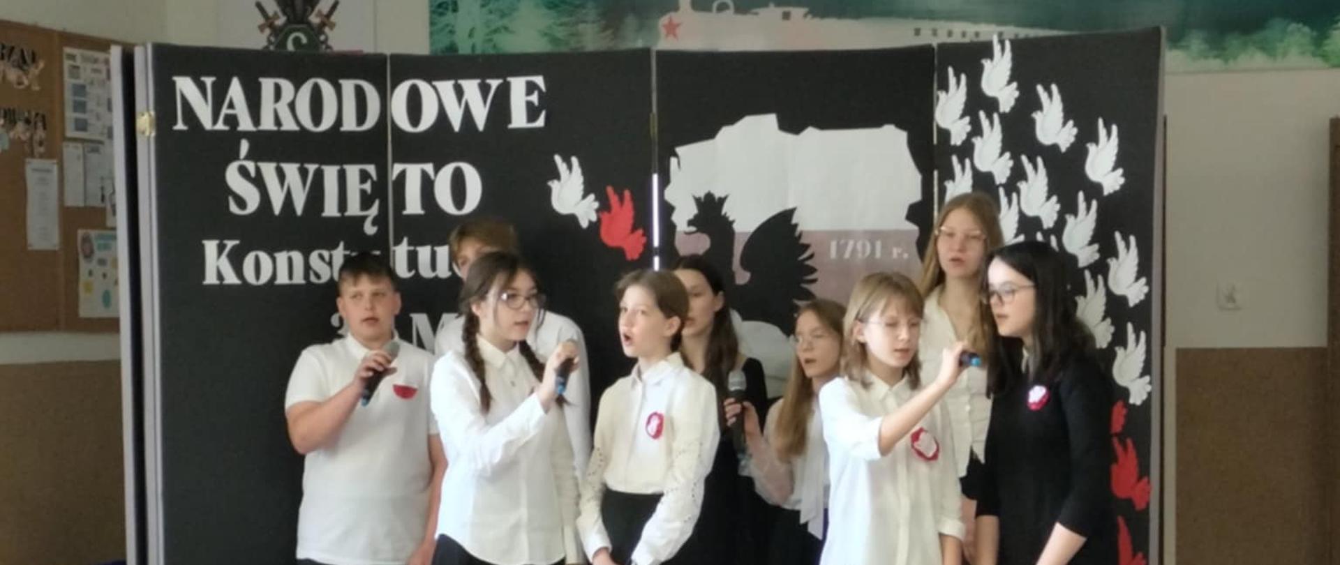 Grupa uczniów ubranych w stroje galowe śpiewa razem piosenkę. Za uczniami znajduje się dekoracja z biało-czerwonymi gołębiami, mapą Polski i napisem: NARODOWE ŚWIĘTO KONSTYTUCJI 3 MAJA.