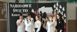 Grupa uczniów ubranych w stroje galowe śpiewa razem piosenkę. Za uczniami znajduje się dekoracja z biało-czerwonymi gołębiami, mapą Polski i napisem: NARODOWE ŚWIĘTO KONSTYTUCJI 3 MAJA.
