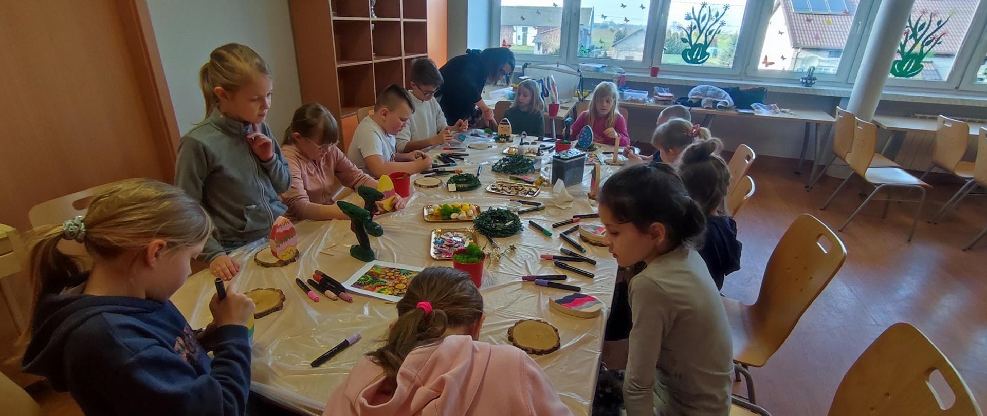 Zdjęcie przedstawia uczestników zajęć którzy dekorują swoje stroiki, malując jajka z drewna flamastrami, w tle Pani prowadząca pomaga uczestnikom w zajęciach