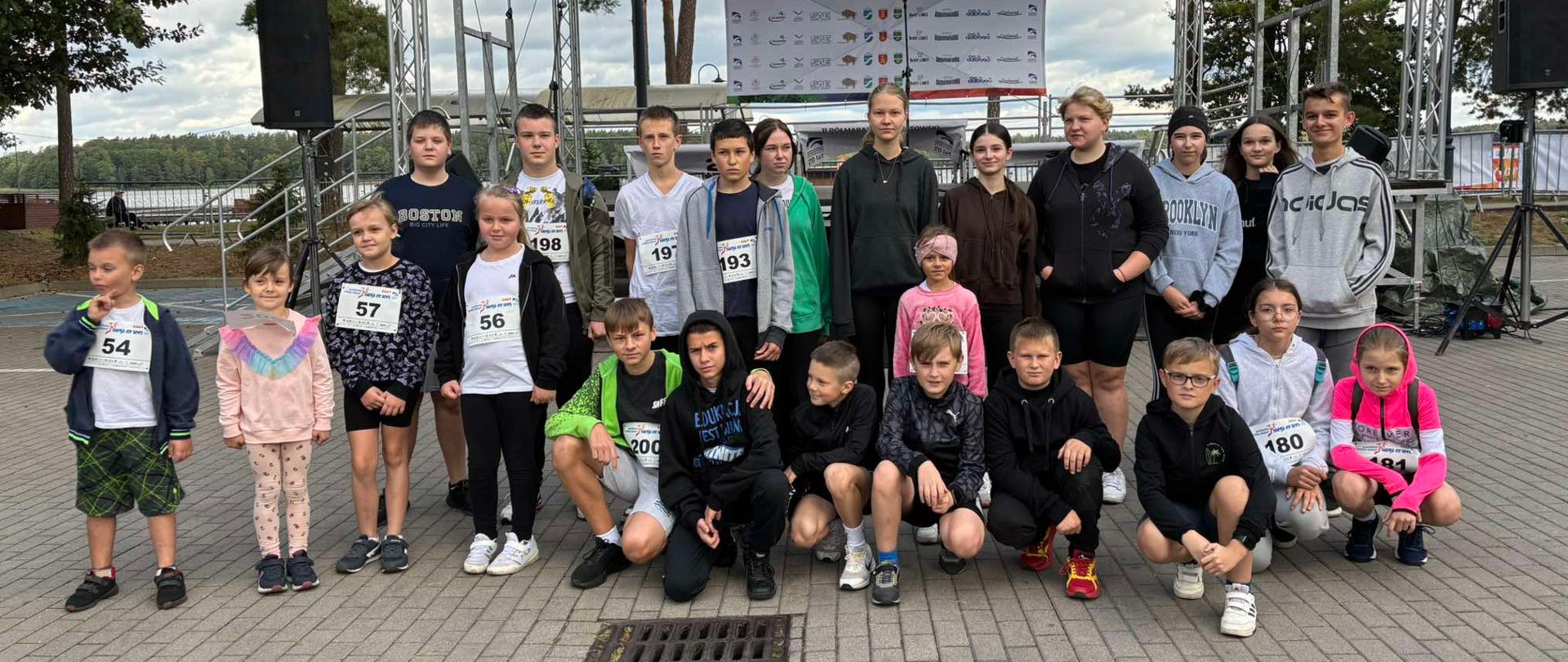 Reprezentanci naszej szkole w Augustowskim Festiwalu Biegowym pozują do wspólnego zdjęcia. Ubrani są w sportowe stroje. Na koszulkach mają naklejone numery startowe.