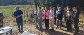 W Lesie Kolanowskim stoi grupa uczniów klasy piątej. Dzieci mają założone na dłoniach rękawice. Obok uczniów stoi mężczyzna i coś mówi.