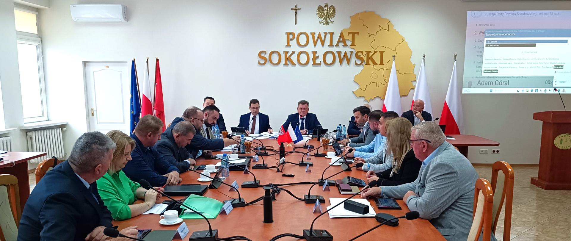 Na zdjęciu widzimy stół w sali konferencyjnej. Po obu jego stronach siedzą naprzeciwko siebie Radni Rady Powiatu Sokołowskiego. W tle ściana, na której zawieszono krzyż oraz godło, i przestrzenny napis Powiat Sokołowski wraz z mapą. Pod ścianą widzimy drugi poprzeczny stół, przy którym siedzi starosta, przewodniczący Rady Powiatu Sokołowskiego wraz z wiceprzewodniczącymi.