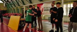 Zdjęcie przedstawia moment wręczania dyplomu jednemu z zawodników przez kierownika Wydziału Edukacji i Sportu Starostwa Powiatowego w Kraśniku panią Agnieszkę Drozd. Po lewej stronie pani Drozd stoi z mikrofonem nauczyciel wychowania fizycznego Zespołu Szkół nr 3 w Kraśniku pan Sławomir Żaba. Podsumowanie turnieju odbywa się na hali sportowej. Z tyłu w tle stoi bramka piłkarska, na której zawieszony jest baner z herbem Powiatu Kraśnickiego i czarnym napisem: Powiat Kraśnicki