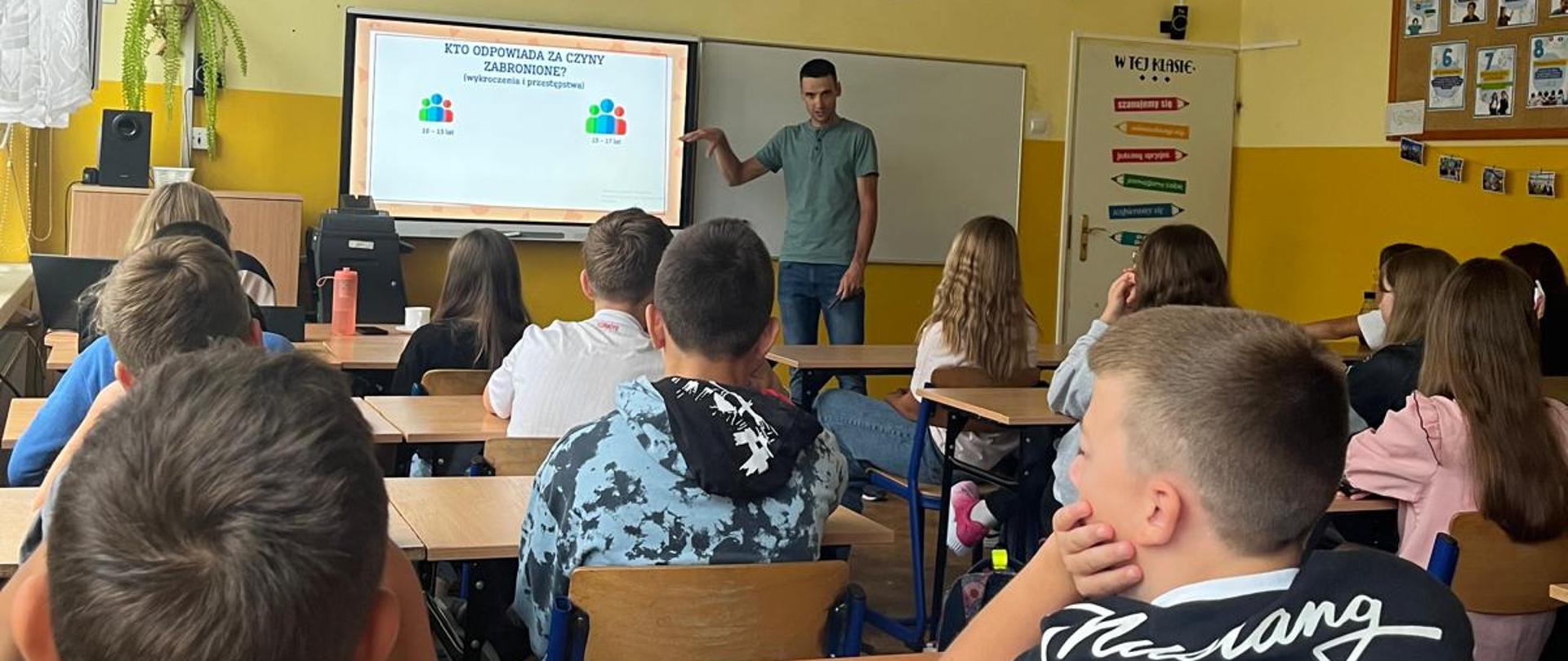 Uczniowie w klasie słuchają nauczyciela stojącego przy tablicy