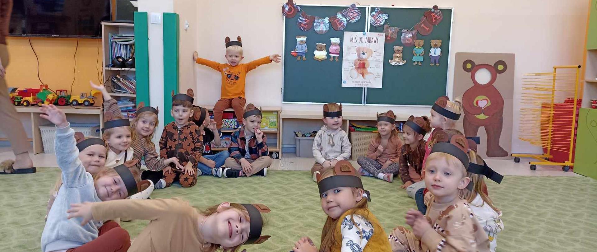 Dzieci siedzą w kole na dywanie , na głowach mają założone opaski z uszkami misia. W tle widać dekorację z okazji Dnia Pluszowego Misia.