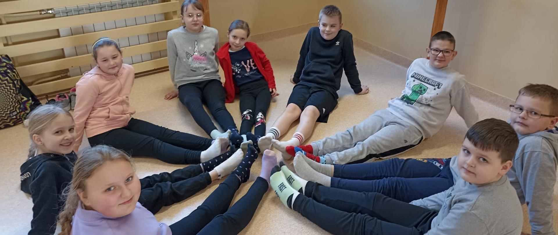 Uczniowie w kolorowych skarpetkach siedzą w kole, z wyprostowanymi nogami, stykają się stopami tworząc w ten sposób kolorowe koło 