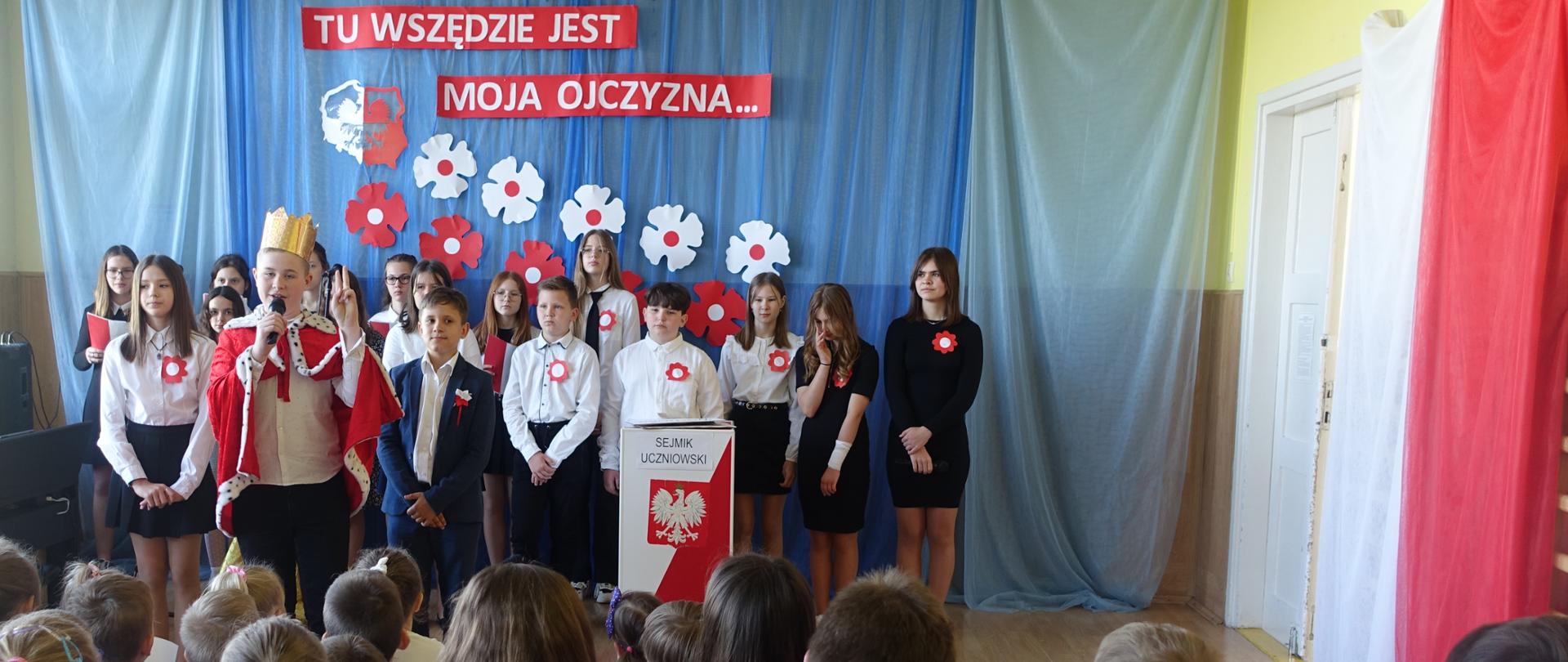 Uczniowie na tle dekoracji z okazji święta Konstytucji 3 Maja.